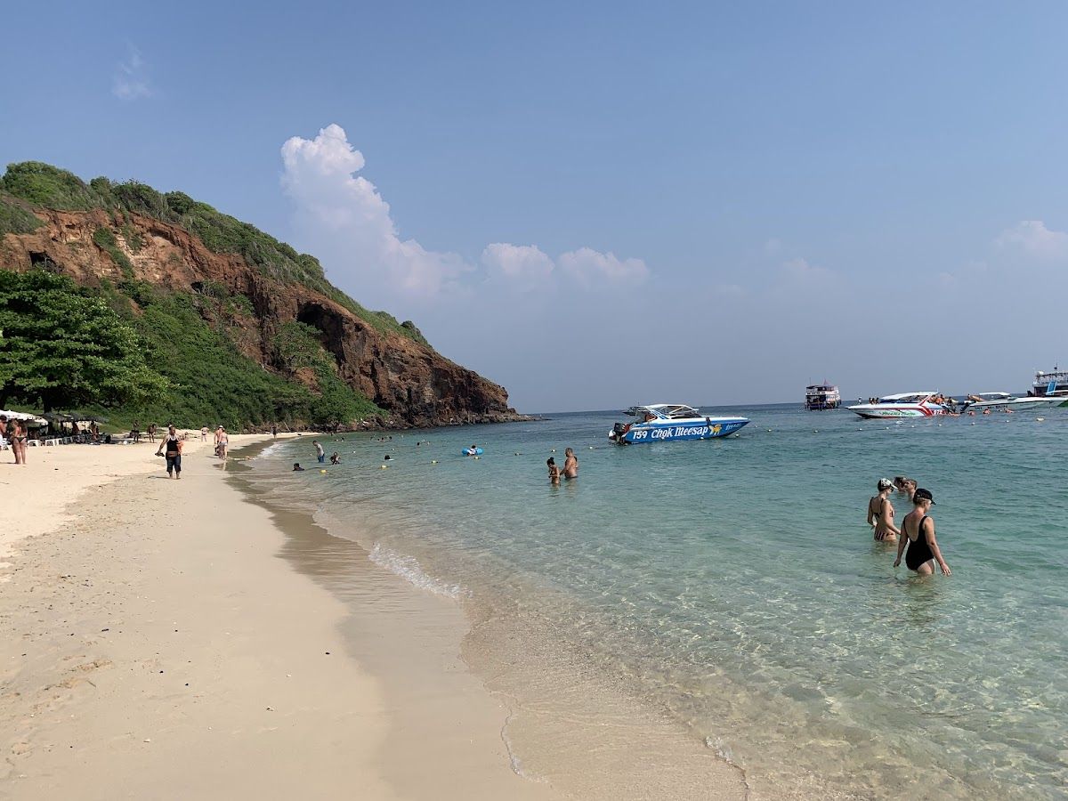Nual Beach on Koh Larn island — clear turquoise water, white sand beach with a dramatic rocky headland, people swimming and speedboats anchored offshore