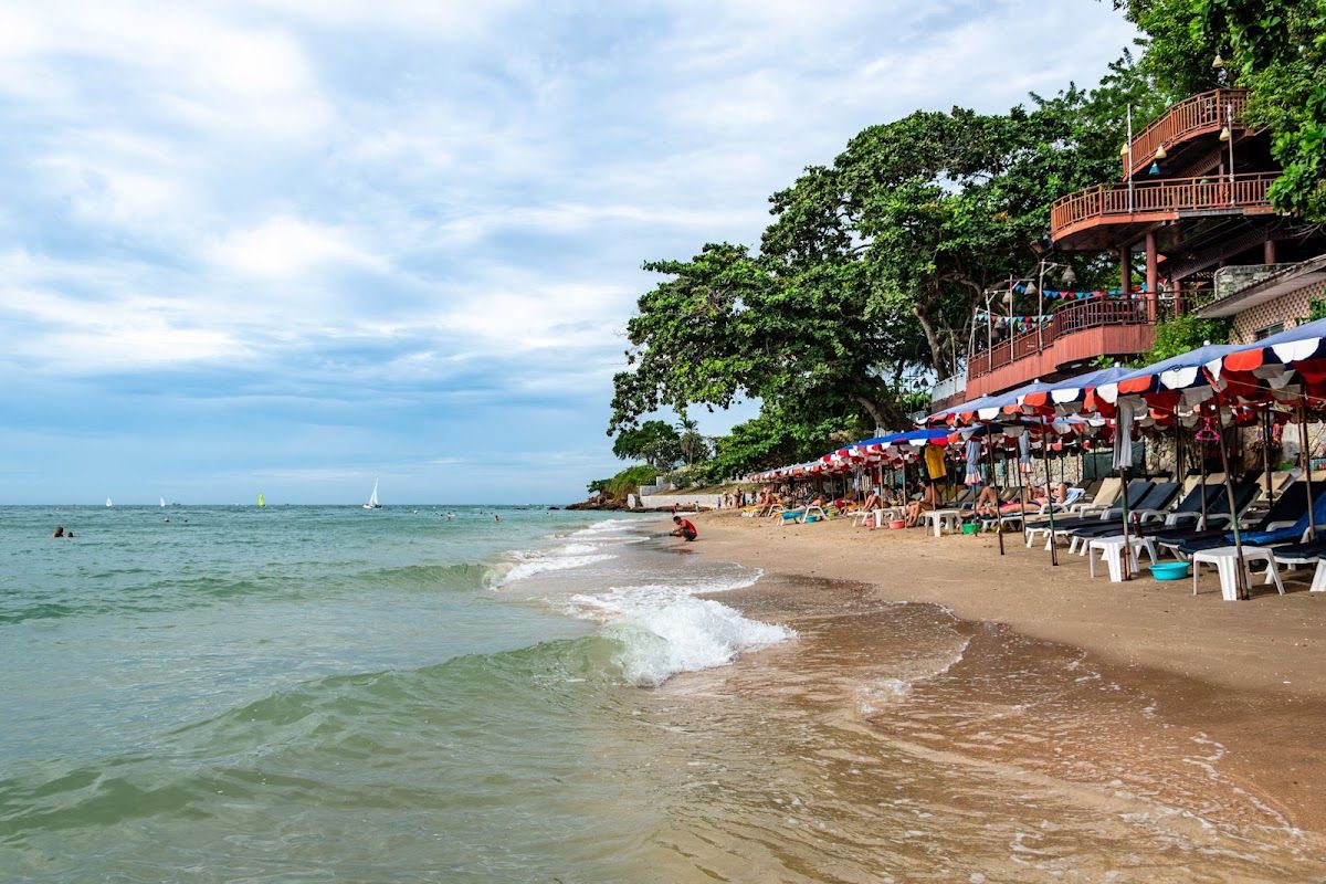 Pratumnak Beach with sunloungers and beach umbrellas along the sand, a beachside restaurant terrace above the shoreline, and sailboats visible on calm water